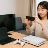自宅のリビングで、テレビ・パソコン・ゲーム機をテーブルに並べ、スマホで撮影している日本人女性。自然光が差し込む明るい室内での作業風景。