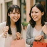 ショッピングバッグを手に笑顔を交わす20代の日本人女性2人。屋外のショッピングエリアで、楽しい時間を過ごしている様子が自然に表現された無料画像